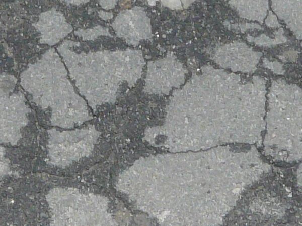 Cracked asphalt texture with many dark, damp patches, and a pothole filled with dark mud and small rocks.